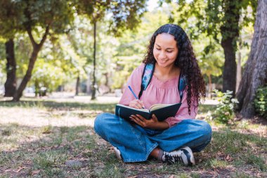 Latin university student woman smiling and writing by hand in her journal, on the grass in a park. High quality photo