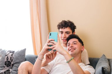Close up of a gay couple lying on the sofa and using the mobile to interact on social media. Daily life. High quality photo