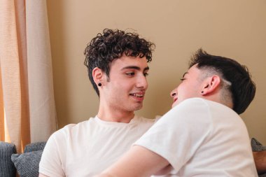 Hugging latin gay couple in pajama on the sofa, taking hands and laughing. Having moment out of routine. High quality photo