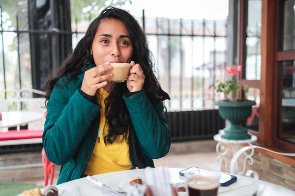 Close up of a young latin american woman, wearing warm clothes, laughing and drinking coffee in a cafeteria. High quality photo