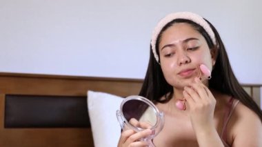 Yanağında gül kuvars yüz silindiri kullanarak yatağında gülümseyen kız. Güzel cilt bakımı. 4k video