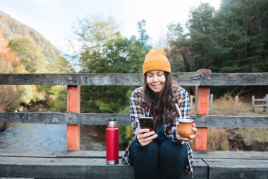 Yerba mate infüzyonunu elinde tutan Latin bir kadının ön görüntüsü ve cep telefonu kırsal kesimde bir nehrin yanında. Uzayı kopyala Yüksek kalite fotoğraf