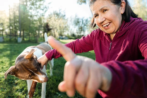 Senior latin woman playing with her young dog puppy and a wooden stick in front of the camera, in the park. High quality photo