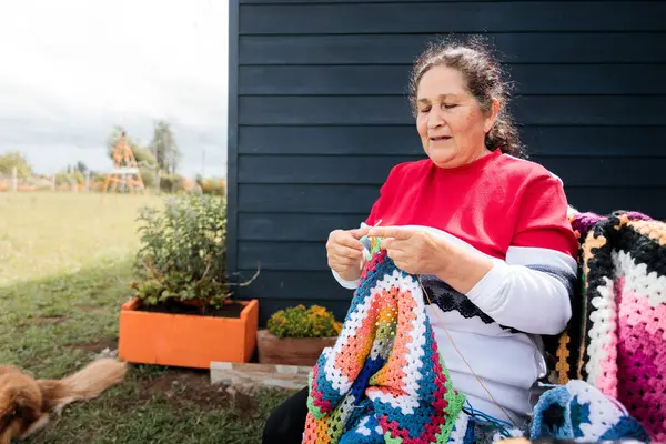 Elderly latin woman with crafts skills embroidering a colorful blanket outside her country house. Low angle view. High quality photo