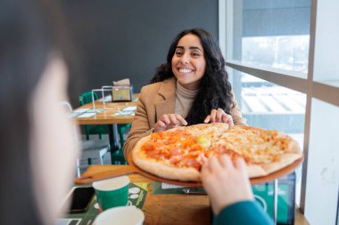 Gülümseyen genç Latin kadın restoranda bir dilim pepperoni pizza yiyor. Bir arkadaşıyla yemeğin tadını çıkarıyor. Yüksek kalite fotoğraf