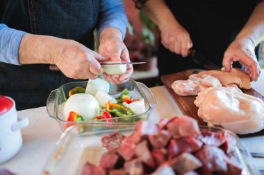 Yaşlı çift beyaz bir masada tavuk ve biftek pişirirken soğan ve biber doğruyor.