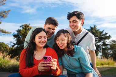 Parkta akıllı telefondan video izlerken gülen dört neşeli Latin öğrenci.