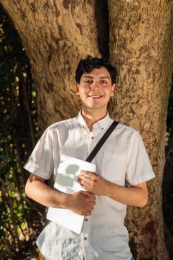 Genç bir erkek öğrenci gülümsüyor, elinde dizüstü bilgisayarla parkta ağaca yaslanıyor, güneşli bir günün tadını çıkarıyor.