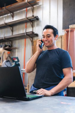 Latin marangoz ve zanaatkar siparişleri dizüstü bilgisayarla idare ediyor ve atölyesinde telefonla konuşuyorlar.