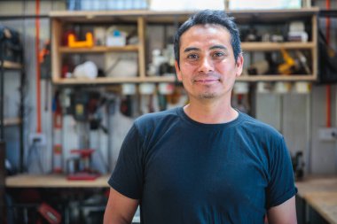 Atölyesinde arka planda aletlerle gülümseyen ve kameraya bakan Latin bir marangoz portresi.