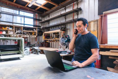 Latin marangoz ve zanaatkar siparişleri dizüstü bilgisayarla idare ediyor ve atölyesinde telefonla konuşuyorlar.