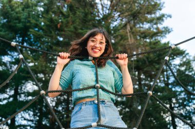 Genç Latin kız güneşli bir günde parkta bir oyun parkında neşeyle halata tırmanıyor, açık hava eğlencesi ve maceranın tadını çıkarıyor.