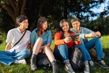 Dört öğrenci parkta çimlerin üzerinde oturmuş tablete bakıyor ve birlikte vakit geçiriyorlar.