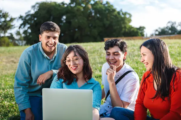 Dört mutlu Latin öğrenci parkta çimlerin üzerinde oturmuş dizüstü bilgisayar izliyor ve gülümsüyor.