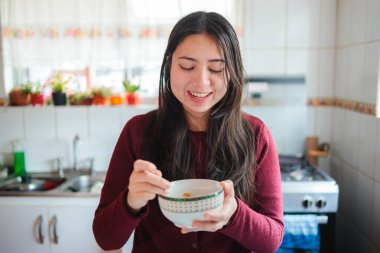Genç bir kadın, neşeli bir mutfakta mısır gevreğinin tadını çıkarırken gülümsüyor. Sahne, sıcak ve davetkar bir atmosferle sakin bir sabah anını yakalıyor.