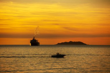 Okyanusun üzerinde gün batımında bir kargo gemisi ve arka ışıkta küçük bir motorlu tekne.