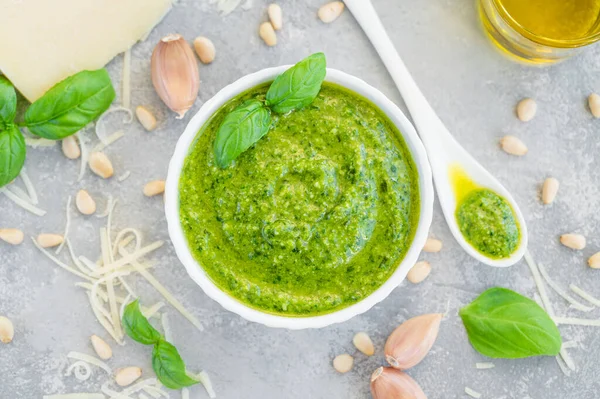 Traditional Italian basil pesto sauce in a white bowl with ingredients for cooking on a gray concrete background. Copy space