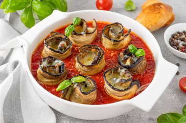 Beyaz seramik kabartma formunda, gri beton zemin üzerinde domates soslu peynirli patlıcan ruloları. Vejetaryen sağlıklı yemek. Seçici odak, üst görünüm