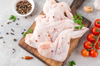 Ahşap kesim tahtasındaki çiğ tavuk kanatları baharatlar, taze otlar, gri beton arka planda domatesler. Kapat.