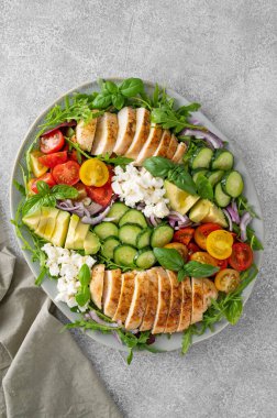 Kızarmış tavuklu salata, avokado, kiraz domatesleri, salatalık, roka peyniri ve gri beton kaplama bir tabakta. Sağlıklı yemek.