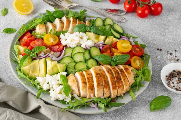 Salad with roasted chicken, avocado, cherry tomatoes, cucumbers, arugula and feta cheese on a plate on a gray concrete background. Healthy food