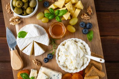 Çeşit çeşit peynirli peynir tabağı, bal, üzüm, fındık, zeytin, yabanmersini ve koyu ahşap arka planda taze otlar. Atıştırmalık şenlik. Üst görünüm, alanı kopyala.