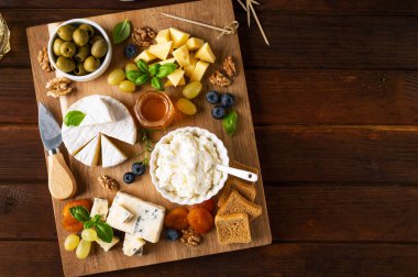 Çeşit çeşit peynirli peynir tabağı, bal, üzüm, fındık, zeytin, yabanmersini ve koyu ahşap arka planda taze otlar. Atıştırmalık şenlik. Üst görünüm, alanı kopyala.