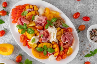 Mozarella soslu Caprese salatası, domates, prosciutto ve gri beton arka planda beyaz bir tabakta ızgara şeftali. Taze yaz salatası. Üst görünüm