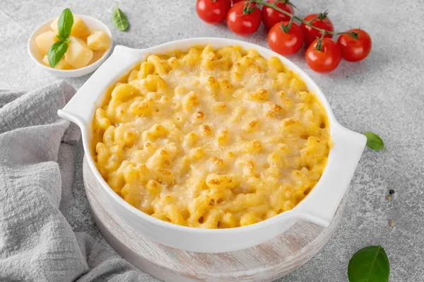 stock image Mac and cheese in a white baking dish on a wooden board on a gray concrete background. American cuisine, comfort food. Copy space