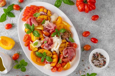 Mozarella soslu Caprese salatası, domates, prosciutto ve gri beton arka planda beyaz bir tabakta ızgara şeftali. Taze yaz salatası. Üst görünüm
