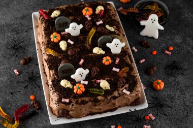 Cadılar Bayramı çikolatalı keki. Çikolata kremalı kek, solucanlar, kafatasları, hayaletler, balkabakları ve şeker parçacıkları ile süslenmiş koyu bir arka planda