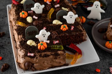 Cadılar Bayramı çikolatalı keki. Çikolata kremalı kek, solucanlar, kafatasları, hayaletler, balkabakları ve şeker parçacıkları ile süslenmiş koyu bir arka planda