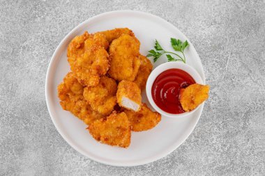 Beyaz tabakta ketçaplı çıtır tavuk külçeleri, gri beton arka planda.