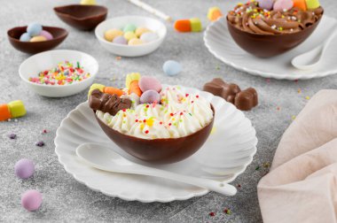 Paskalya tatlısı. Çikolatalı yumurtalar çikolata şekerleri ve şeker parçacıkları ile süslenmiş gri beton arka planda beyaz bir tabakta. Boşluğu kopyala