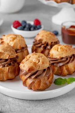 Çikolata kremalı profiteroller, taze böğürtlen ve gri beton arka planda bir tabakta çikolata. Lezzetli bir Fransız tatlısı