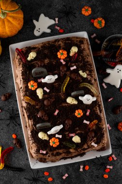 Cadılar Bayramı çikolatalı keki. Çikolata kremalı kek, solucanlar, kafatasları, hayaletler, balkabakları ve şeker parçacıkları ile süslenmiş koyu bir arka planda