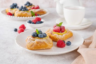 Danish puff pastry cakes with custard cream and fresh berries, sprinkled with powdered sugar on a white plate on a concrete background