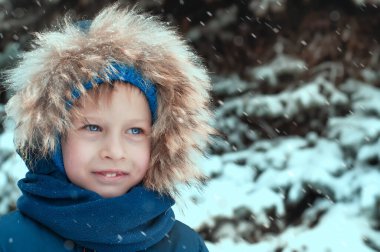 Kış kıyafetleri giymiş bir çocuk kar kaplı bir Noel ağacının arka planına gülümsüyor, kar yağıyor. Yüksek kalite fotoğraf