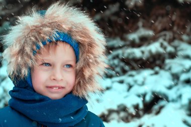 Kış kıyafetleri giymiş bir çocuk kar kaplı bir Noel ağacının arka planına gülümsüyor, kar yağıyor. Yüksek kalite fotoğraf