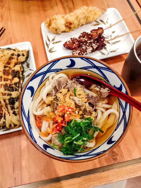 stock image udon noodles served with chicken satay and seaweed tempura, accompanied by a glass of iced lemon tea. This traditional Asian dish combines the flavors of Japanese and Indonesian cuisines