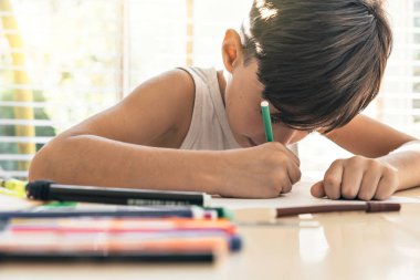 Latin çocuk (7-8) deftere çizim yapıyor. Görüntünün arka planındaki parlak ışık güvenli ve besleyici bir ortamın çocukların gelişmesini ve ifade özgürlüğünü teşvik ettiğini gösteriyor..