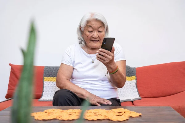 Older woman of Japanese ethnicity with grey hair looking at her mobile phone screen, sitting at home. Online communication using a smartphone.