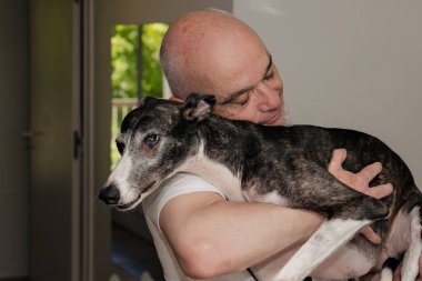 Yetişkin bir adamın portresi. Evde bir tazı köpeğini kucaklıyor. Sevecen bir jesti paylaşıyor..