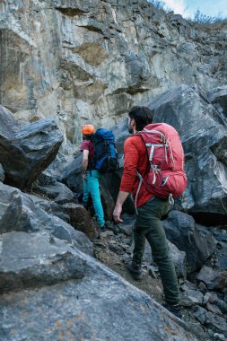Sırt çantalı iki yürüyüşçü zorlu bir tırmanışa hazırlanan kayalık bir uçuruma yaklaşıyor. Kask ve tırmanma ekipmanları takıyorlar. Açık hava macerasına hazırlar..