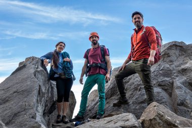 Bir grup farklı dağcı nefes kesen bir dağın zirvesinde zaferi kutluyor, kameraya neşe ve başarıyla bakıyor. Gökyüzünün altında muhteşem bir manzara..