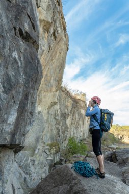 Uçurumun kenarında duran cesur kadın yürüyüşçü, güvenlik kaskını kararlılıkla ayarlıyor. Arka planda bulutsuz bir gökyüzünün altında görkemli bir dağ manzarası.
