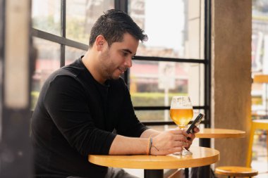 Latin bir adamın gün içinde barda akıllı telefonuyla mesajlaşırken bir bardak biranın keyfini çıkardığı profil. Rahat gündüz atmosferi ve modern bar ortamı gündelik ve keyifli bir anı geliştirir..