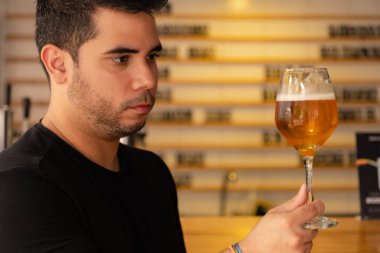 Genç bir Latin adam bir barda el yapımı biranın tadını çıkarıyor. Tadına baktığı biranın rengini dikkatle inceliyor ve bira kültürüne olan minnettarlığını gösteriyor..