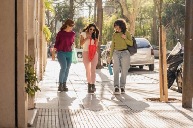 Üç kadın güneşli bir caddede yürüyor, sohbet ediyor ve gülümsüyor ve tekrar kullanılabilir çantalar taşıyor. Rahatlamış atmosfer, canlı bir şehir ortamında arkadaşlığın ve açık hava eğlencesinin altını çiziyor..