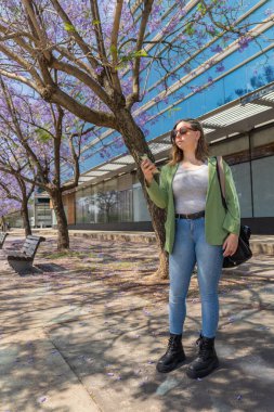 Şehir parkında telefonunu kontrol eden genç, profesyonel bir kadın. Çiçek açmış Jacaranda ağaçları var.